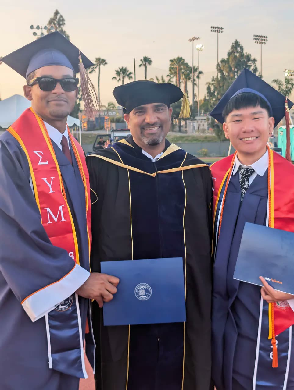 Two graduating members in Sigma Upsilon Mu stoles with a faculty advisor at commencement
